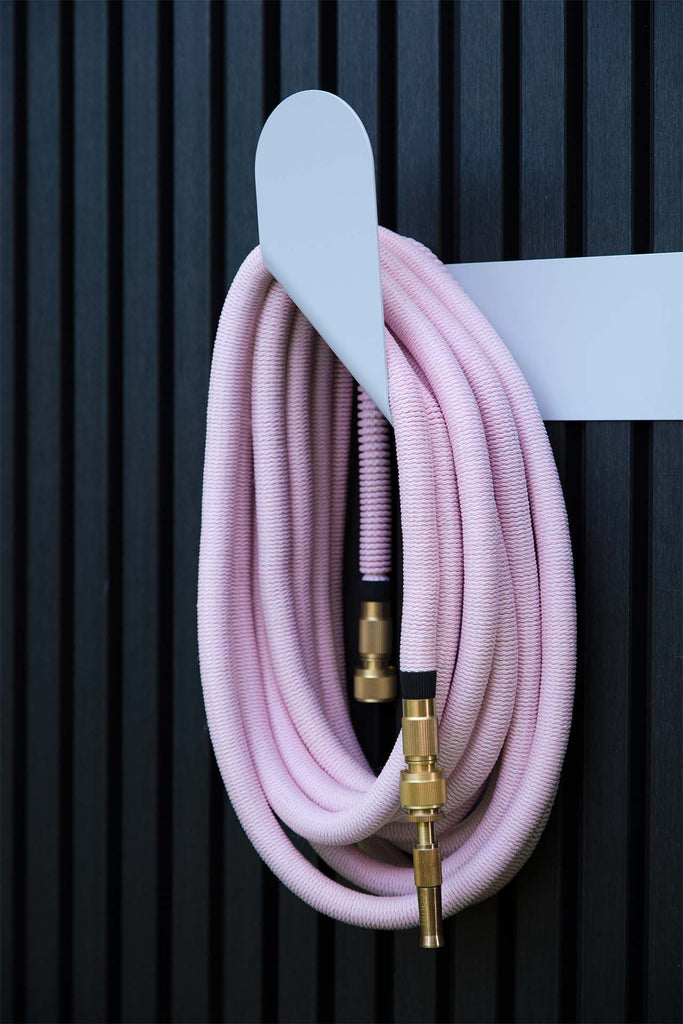 Fritz Tango Soft Expandable Garden Hose in pink, hanging gracefully on the sleek white Hendrix Hose Holder against a modern black panel wall. A perfect blend of function and style.