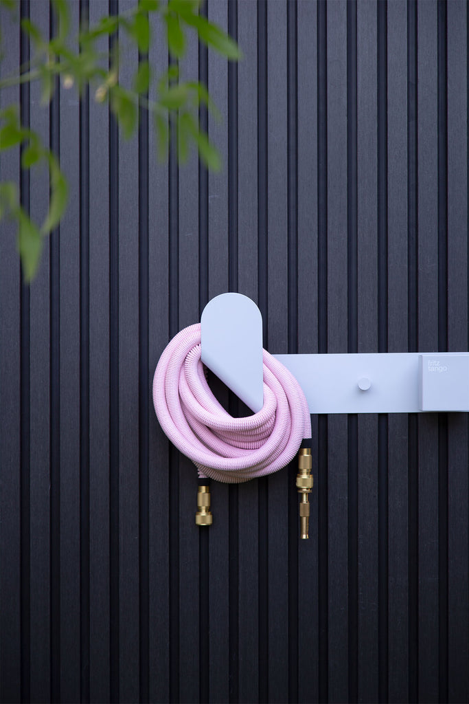 Fritz Tango Soft Expandable Garden Hose in pink, neatly coiled on the sleek white Hendrix Hose Holder against a textured black panel wall. A modern and practical garden accessory.