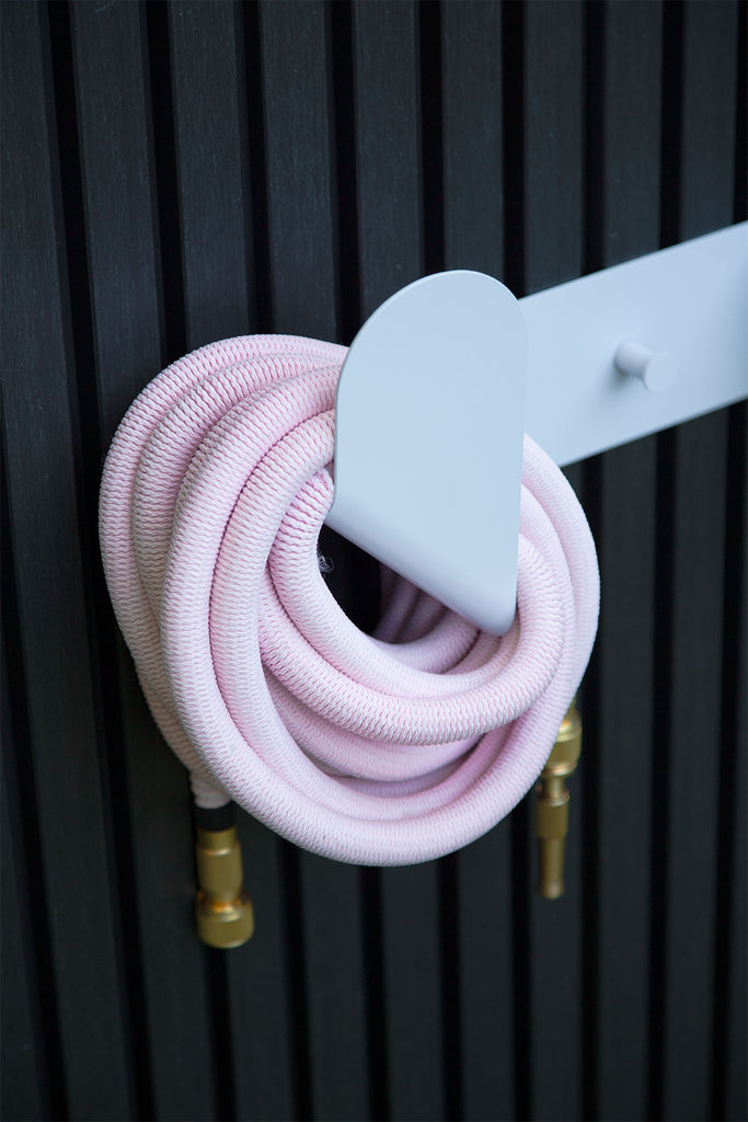 Fritz Tango Soft Expandable Garden Hose in pink, neatly coiled on the minimalist Hendrix Hose Holder against a sleek black panel wall. Perfect for organized and stylish garden storage.