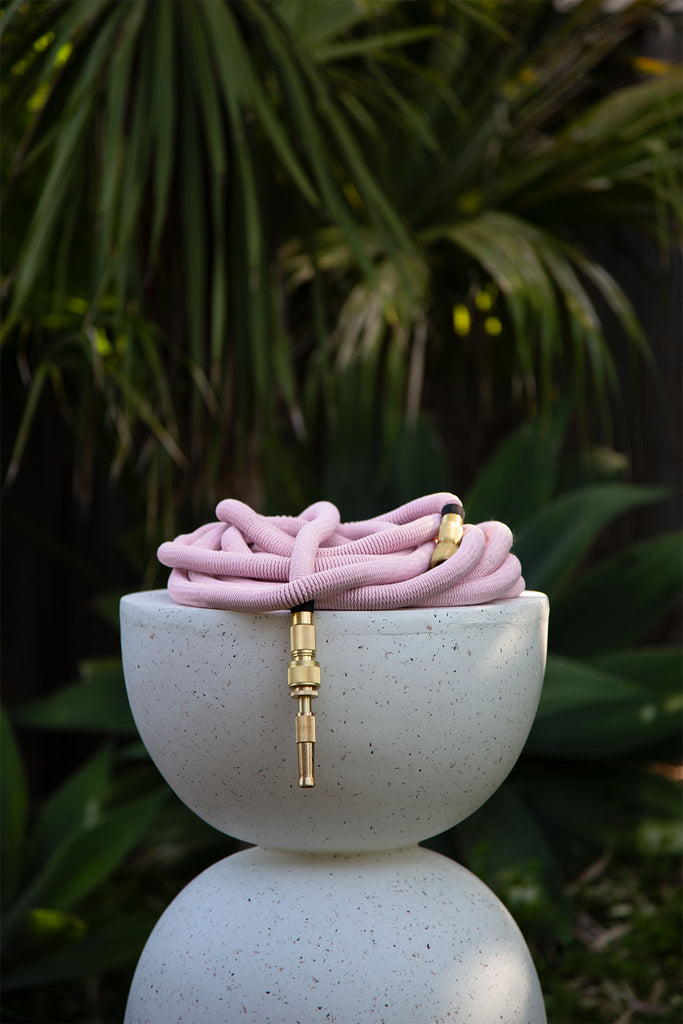 Fritz Tango Soft Expandable Garden Hose in pink, elegantly coiled on a modern speckled white designer stool with lush greenery in the background. Durable, lightweight, and stylish for garden enthusiasts.