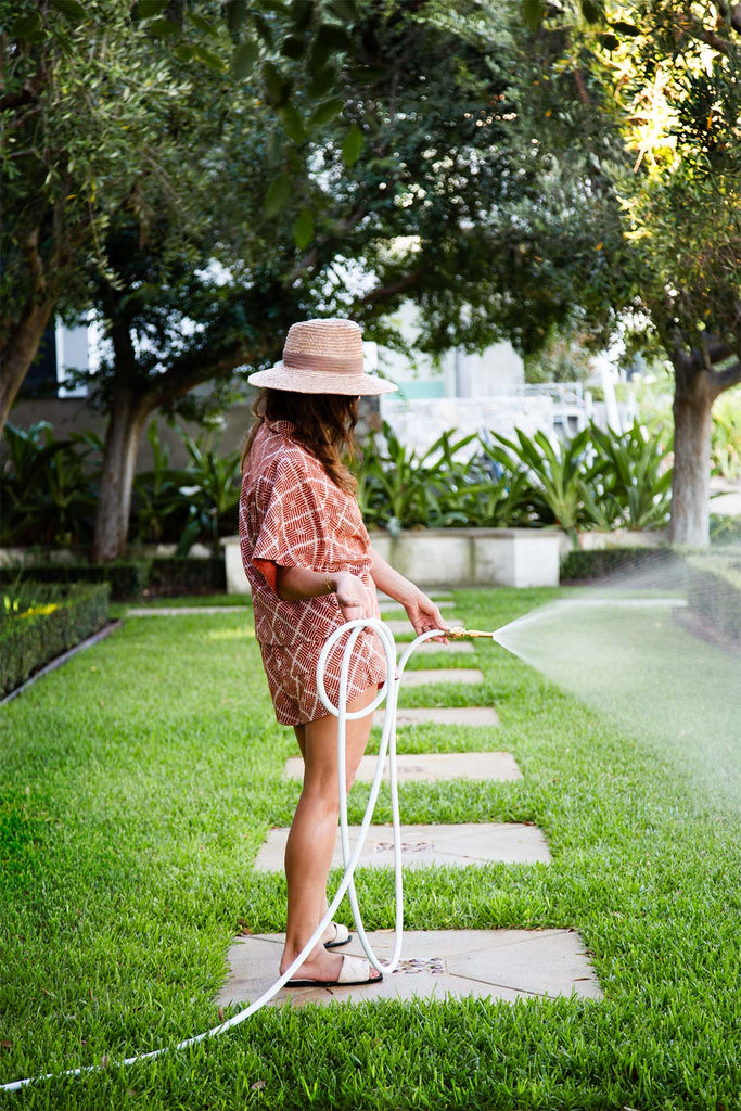 Woman watering a green lawn with a white flexible garden hose, wearing a patterned summer outfit and straw hat. A tranquil outdoor scene with lush foliage.